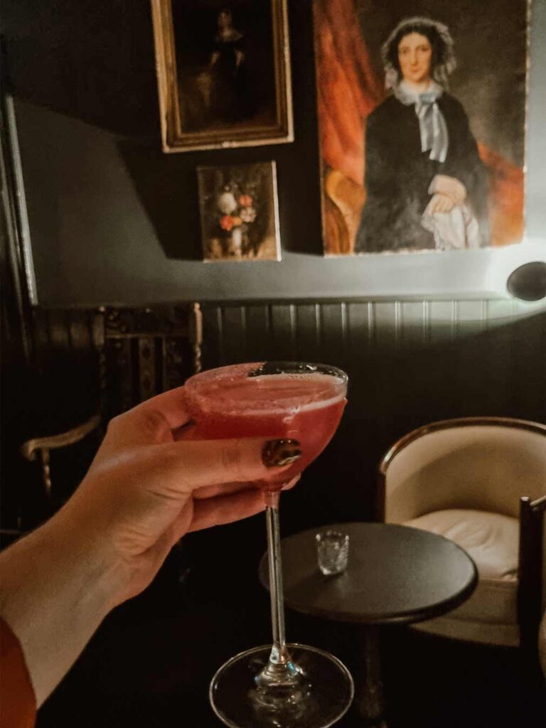 A woman holding a tasty pink cocktail at Pulitzer's Bar. You can see old paintings and portraits in the background and the bar is in a low lit setting. 