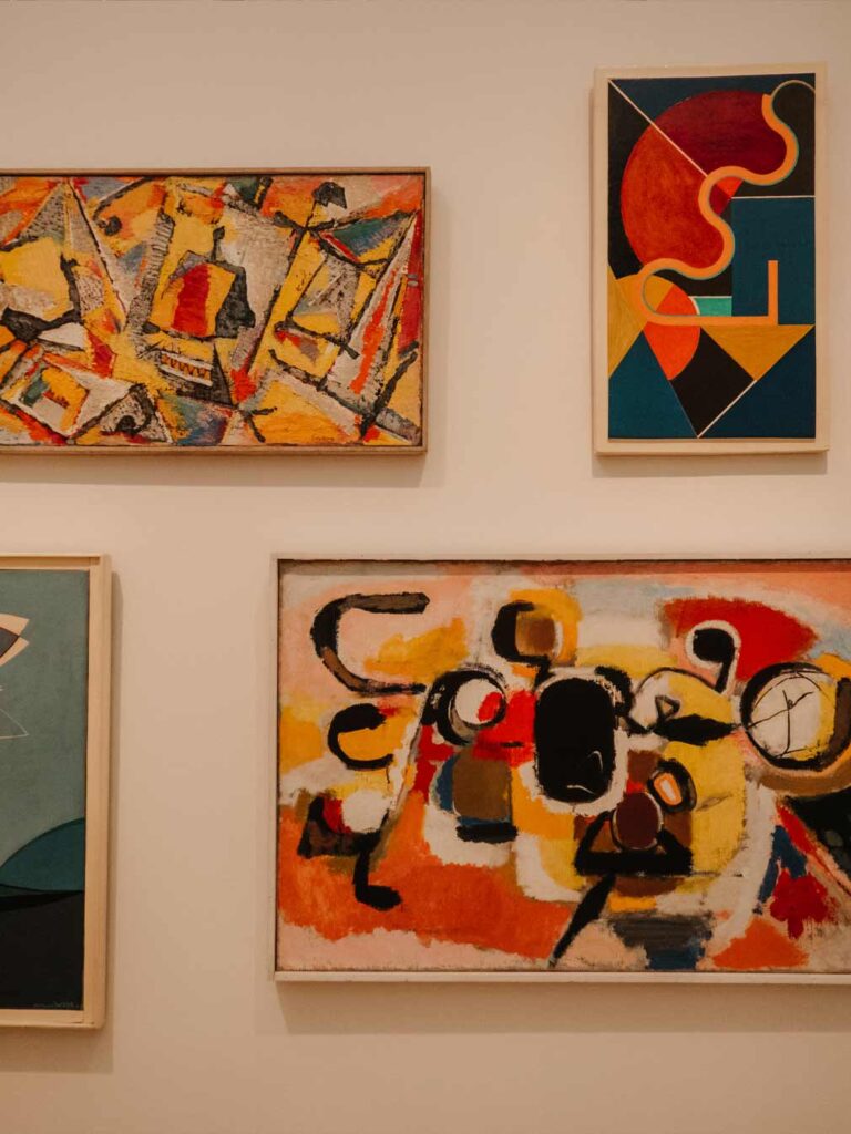 Colourful art on display at Stedelijk Museum. The colours used are mostly black, white, red, blue, yellow and a little bit of green. 

The art is in quite a geometric abstract style painted with oil paints and held inside wooden frames.