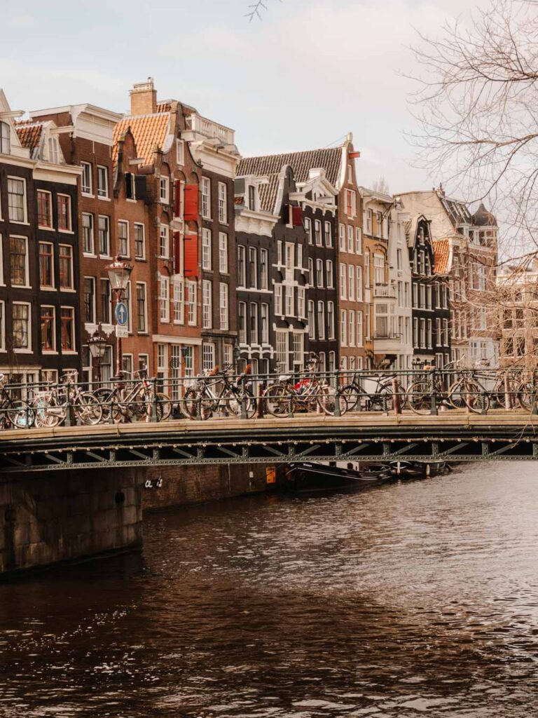 Views of traditional crooked buildings and canals in Amsterdam. Some of the buildings have wooden shutters on the windows and the buildings have been build with different coloured bricks such as light brown, dark brown and black.