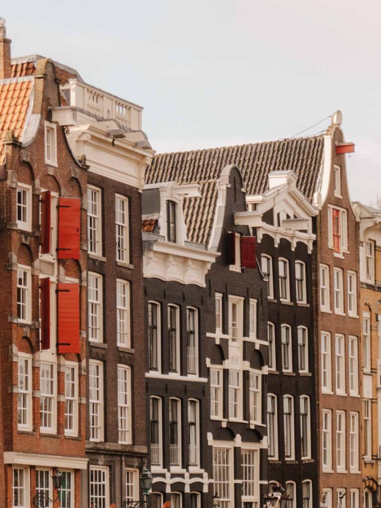 Crooked buildings in Amsterdam. The buildings stand at different heights and have been built with either light brown, dark brown or black coloured bricks. There are red shutters on some of the buildings and some also have hooks on the top - many buildings across the city have these hooks.
