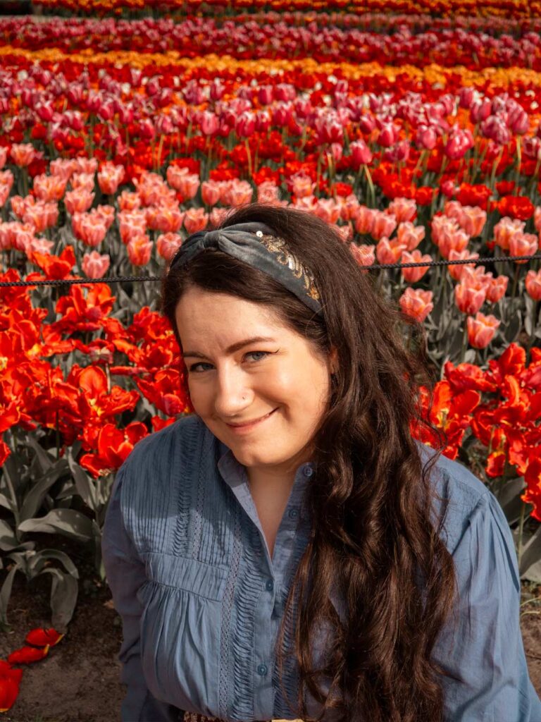 A brunette woman at Keukenhof tulip gardens with a colourful patch of tulips behind her. The tulips are a variety of colours including red, pink and yellow.