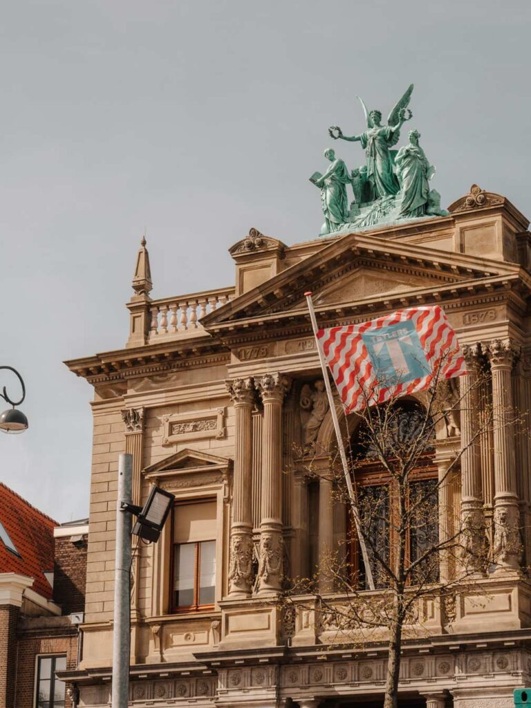 The exerior of the Teylers Museum in the Netherlands. It has a grand facade with a statue of an angel and two peole on the top. It also has a flag with the museums logo that says 'Teylers' on it. The flag is decorated with a red and white wavy pattern and the logo is blue and white.