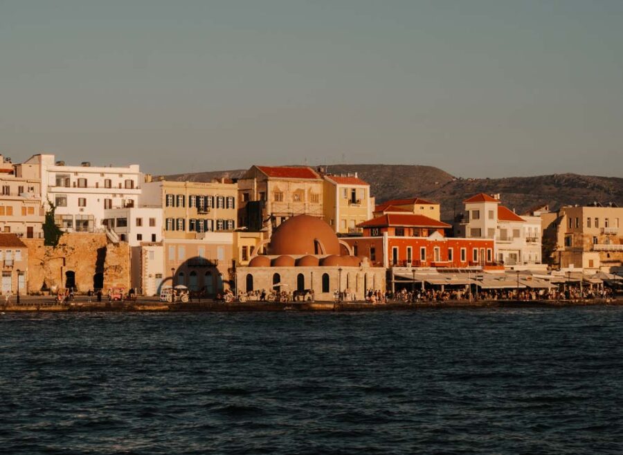 Views of the old Venetian Harbour and the Kucuk Hasan Mosque in Chania. The harbour is full of colourful buildings and you can see the sea in the foreground. The mosque has a large dome that's been painted a dark pink colour.