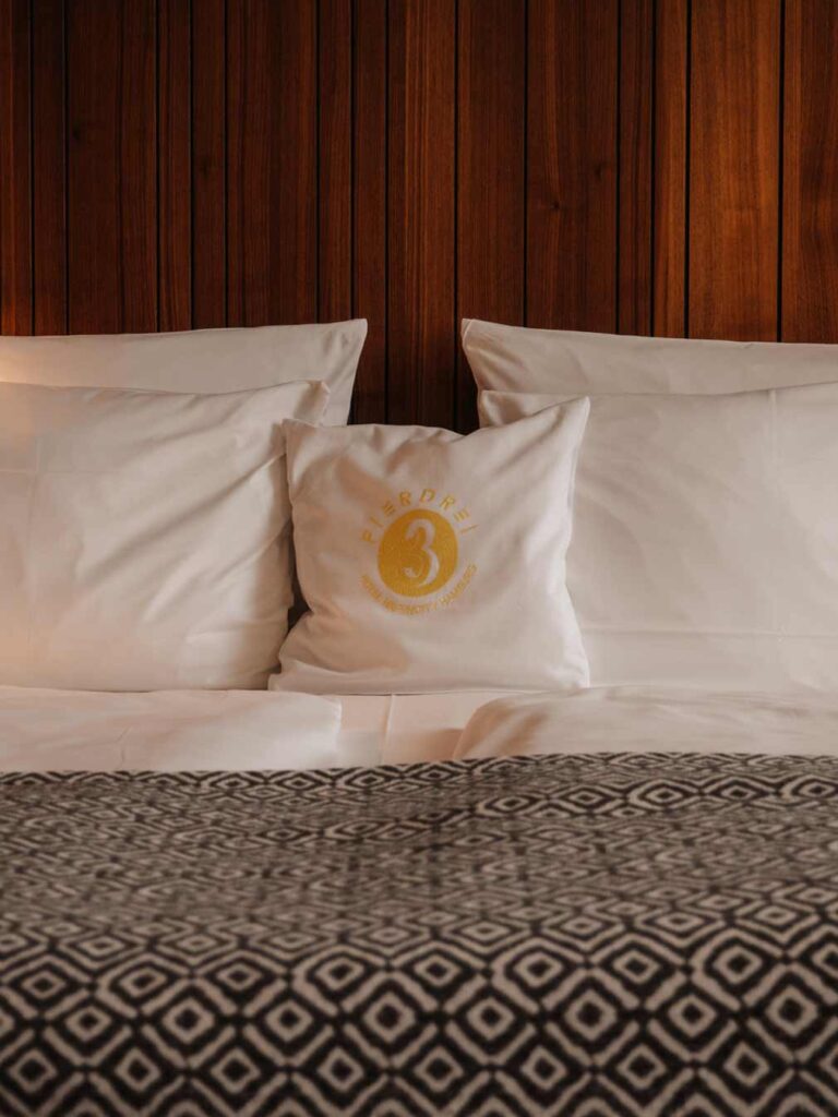 The bed in the medium plus room which is decorated with plain white bedding, a cushion with the hotel's name on it and a grey geometric woven blanket. To the right there are magazines hanging on a rack and the feature wall is a walnut wood style.