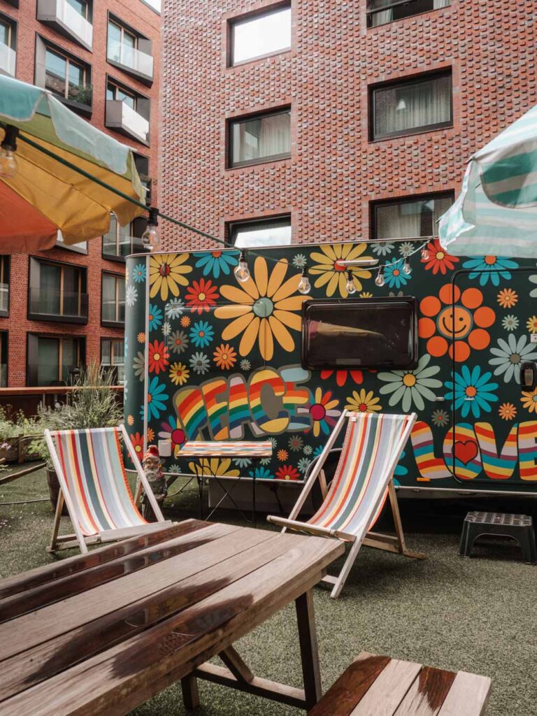Flower Power Camper at Camper City. The campervan is decorated with brightly coloured flowers all over and says 'Peace and Love' in a rainbow of colours. There are also some multi-coloured striped deck chairs, a bench and some sun umbrellas (despite it being a rainy day).