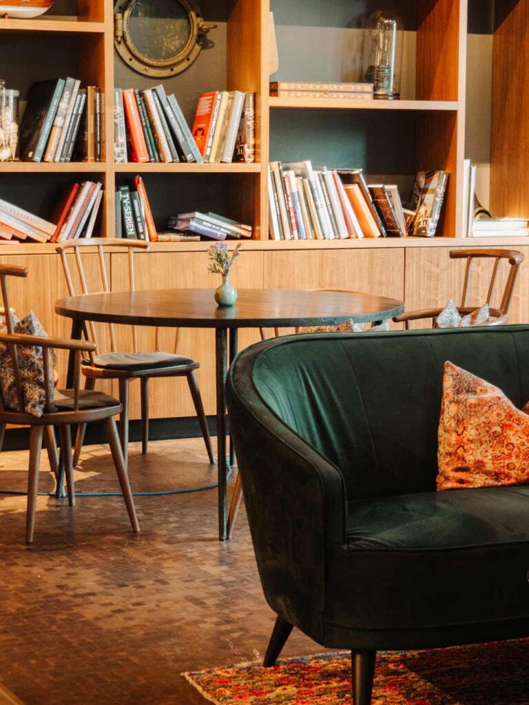 Guest lounge at Pierdrei Hotel Hafencity with lots of books on display, cost seating areas and a wooden table and chairs.