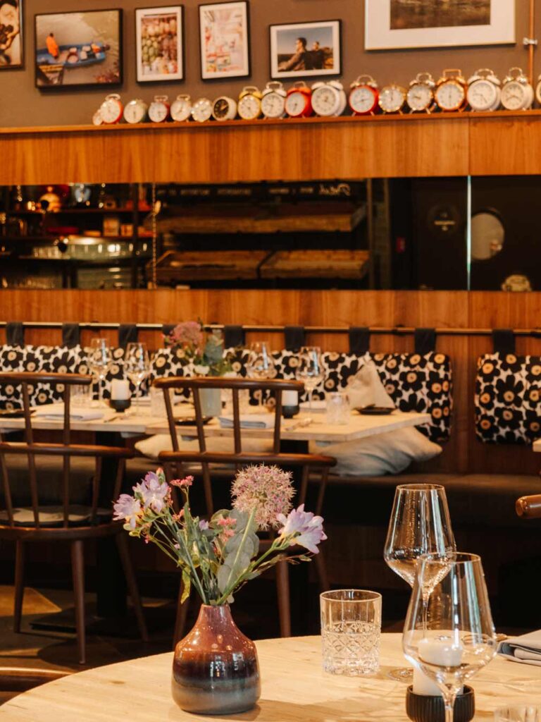 The Kitchens Restaurant at Pierdrei Hotel Hafencity. There is a light table in the foreground that holds a small vase of flowers and some glasses. In the distance, you can see more tables set for dinner and Marimekko printed back rests. There is also a row of different coloured alarm clocks sat on the wooden display shelf and a gallery wall.