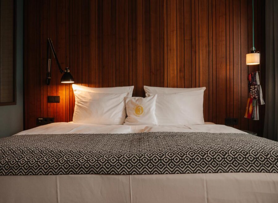The bed in the medium plus room which is decorated with plain white bedding, a cushion with the hotel's name on it and a grey geometric woven blanket. To the right there are magazines hanging on a rack and the feature wall is a walnut wood style.