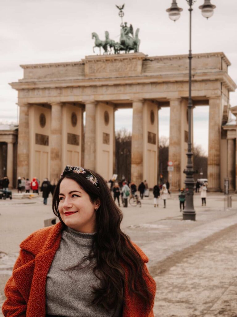 Lauren (the owner of this blog) in front of the Brandenburg Gate in Berlin, Germany. Lauren is wearing an orange coat, grey jumper and black headband. In the background, you can see some other tourists and the slightly snowy pavement as she was visiting in January.