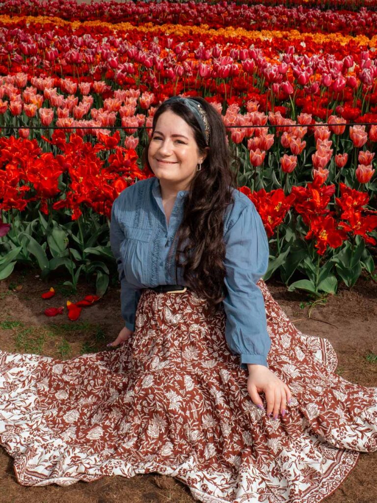 Lauren sat in front of rows and rows of colourful tulips at Keukenhof. She's wearing a red and white patterned maxi skirt, a blue shirt and a blue sequined headband.