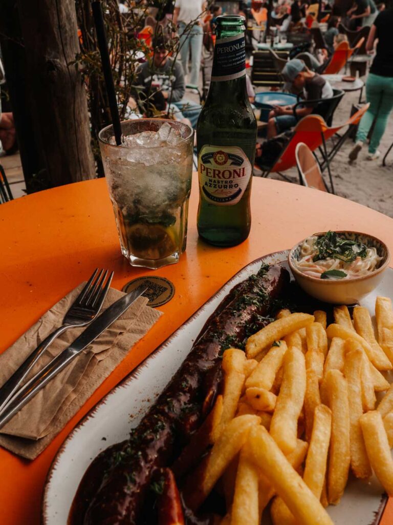A plate of currywurst, chips and coselaw at Stand Pauli. There is also a mojito cocktail and a bottle of Peroni beer on the round orange table.