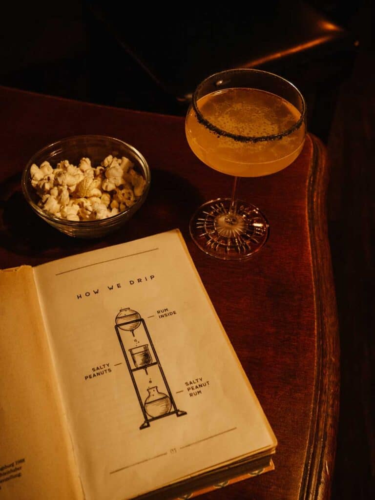 A cozy scene featuring a book on a table, paired with a cocktail and popcorn. The book explains how the cocktails are made at Drip Bar with an illustration.