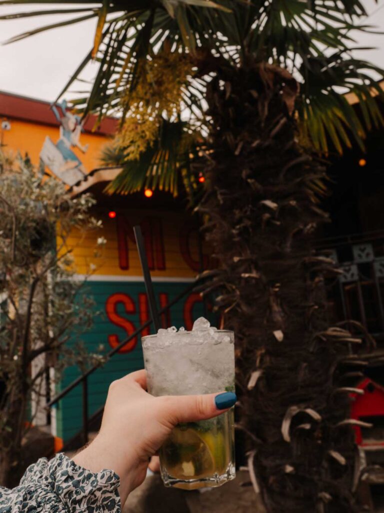 Lauren (the owner of this blog) holding a mojito cocktail at Stand Pauli. You can also see colourfully painted walls and palm trees in the background.