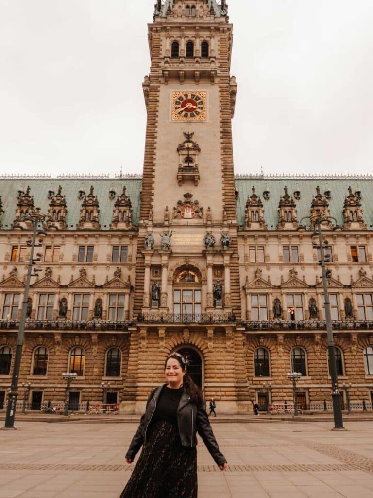 Lauren (the owner of this blog) posing outside the City Hall in Hamburg. Lauren is wearing a black tshirt, black floral skirt and a black leather jacket. The City Hall is an impressived neo-renaissance building with a green roof, prominent clock face and an elegant design.