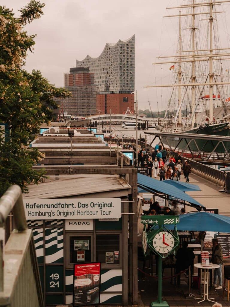 Views of St. Pauli Pier on an overcast and rainy day. You can see ticket offices for port cruises, Rickmer Rickmers Museum and people walking along the waterway. You can also see the striking Elbphilharmonie in the far distance.