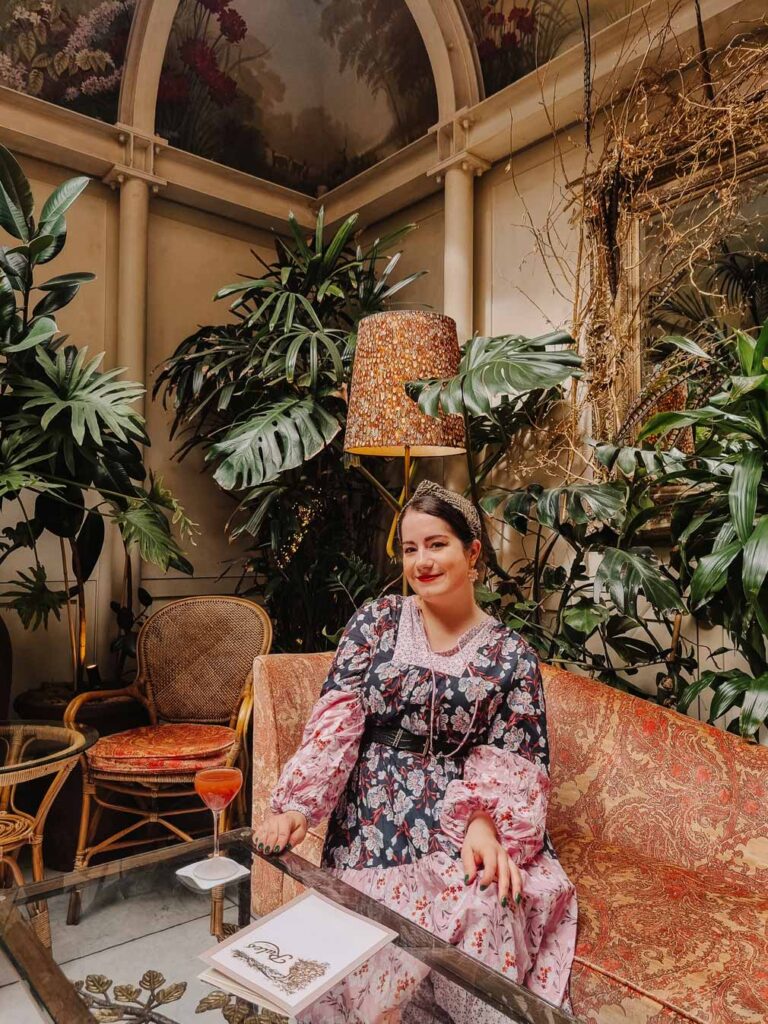 Lauren enjoying cocktails at Upstairs at Rules near Covent Garden. She is sat on a paisley printed sofa surrounded by plants and painted ceilings.