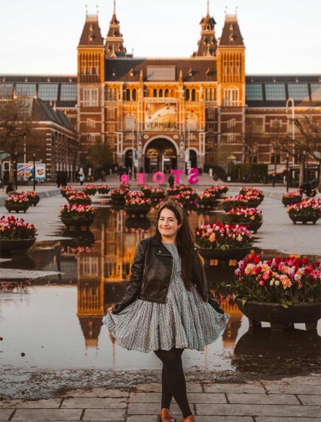 Lauren (the owner of this blog) in front of the Rijksmuseum. There is a large puddle with a reflection of the museum behind her as well as plant pots with loads of colourful tulips in them. Lauren is wearing a blue and white floral dress, with tights and brown shoes.
